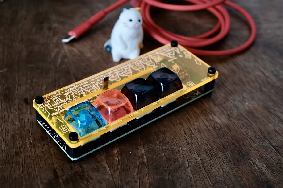 A Disarray Desnarler in a yellow acrylic case with blue, pink and two smoky keycaps, next to a cat figurine and a red USB cable.
