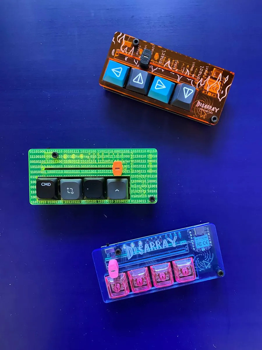 Three Disarray Desnarler macropads, each with four keys and a fader, in orange, neon green and translucent blue acrylic cases.