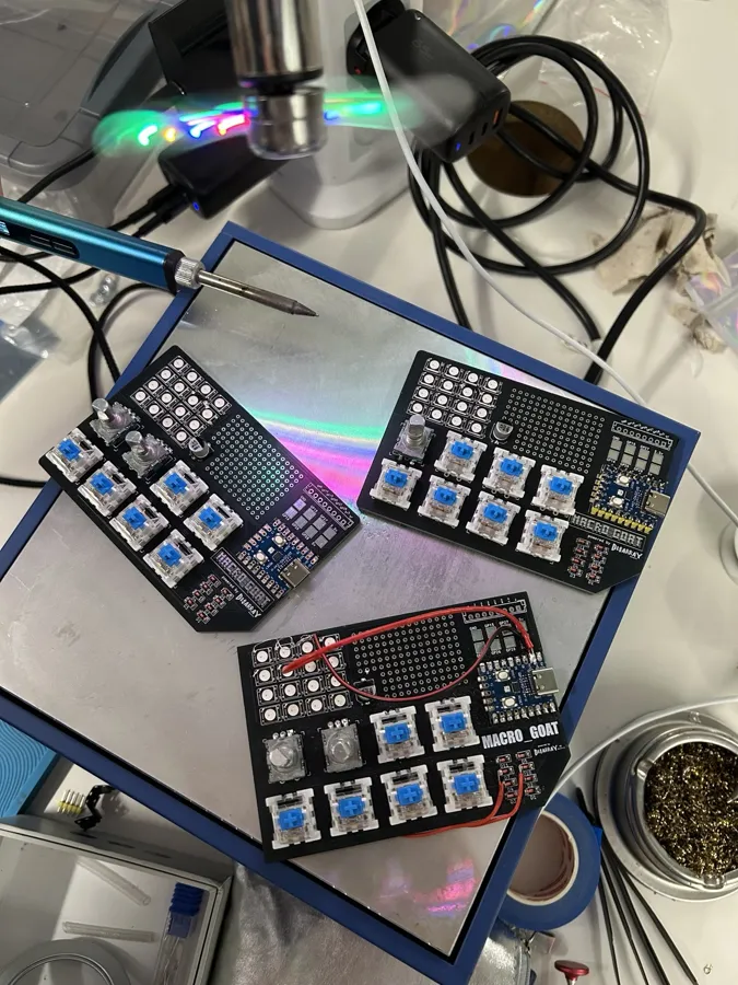 Three assembled macro_GOAT boards with blue key switches and rotary encoders on a workbench next to a soldering iron.