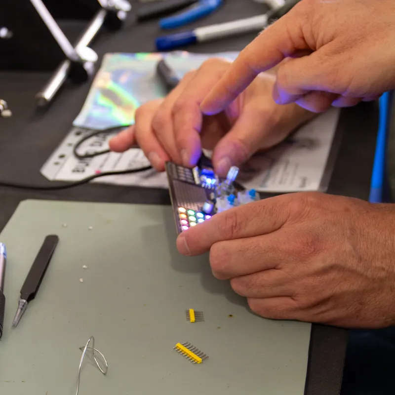 Hands holding a freshly soldered macro_GOAT with its LED grid lit up, next to a soldering iron and tweezers on a workshop mat.
