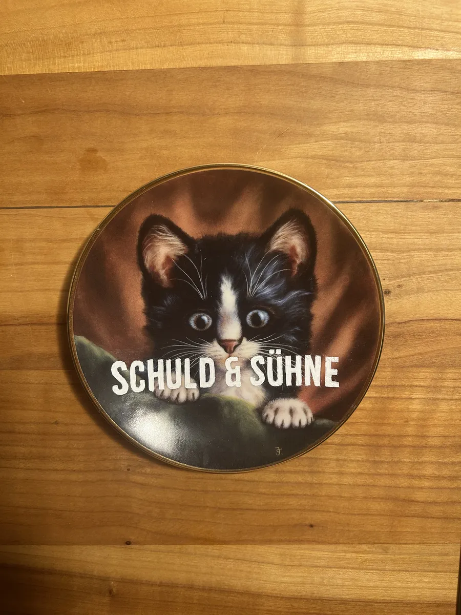 A small plate with a wide-eyed black and white kitten, lettered Schuld & Sühne in white.